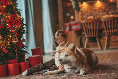 Candid authentic happy little boy in knitted beige sweater hugs dog with bow tie at home on xmas