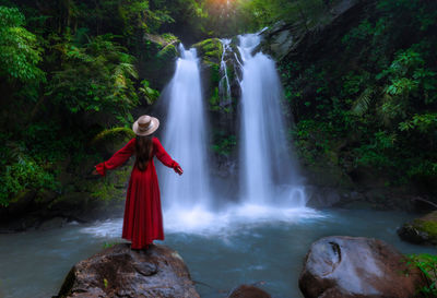 Rear view of woman standing on rock against waterfall in forest