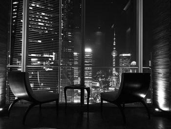 Empty chairs by window against illuminated skyscrapers