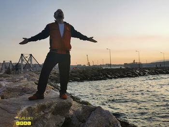 Man standing on rock by sea against sky during sunset