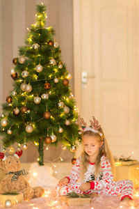 Full length of cute girl sitting by illuminated christmas tree