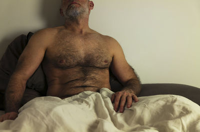 Portrait of shirtless adult man lying on bed against white wall