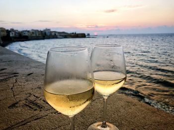 Close-up of wineglasses on retaining wall by sea against sky during sunset