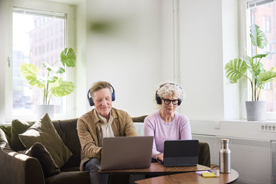 Senior man and woman sitting in living room and using laptop and digital tablet to edit podcast