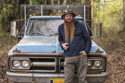 Portrait of confident bearded man standing against old pick-up truck in forest