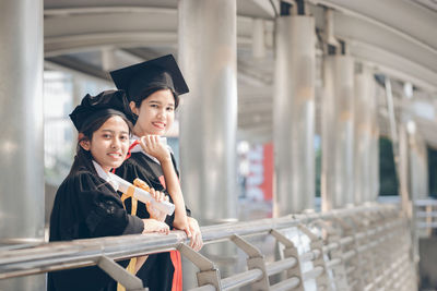 Portrait of friends wearing graduation gown while standing by railing