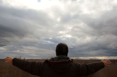 Rear view of mature man with arms outstretched standing against cloudy sky