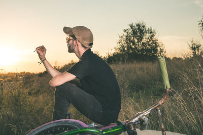 Man sitting with bicycle on field against sky during sunset