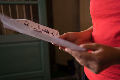 Cropped unrecognizable female in red t shirt examining manual paper while standing in cozy room at home