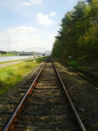 Railroad track with trees by riverbank against cloudy sky