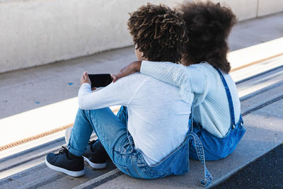 Back view of unrecognizable curly haired african american teen siblings in similar denim clothes hugging and browsing smartphone while sitting together on street