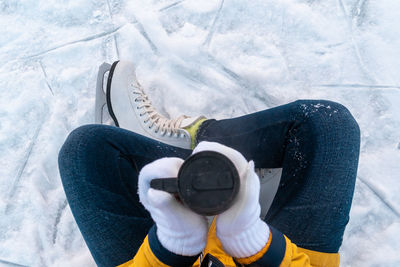 Young woman in ice skates sitting on the snow and holding mug with coffee. close up of female hands.