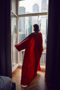 Woman in a red bathrobe stands at a large window in an apartment opposite a skyscraper