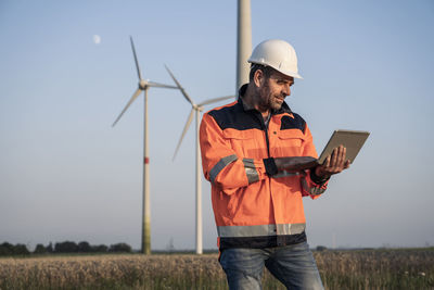 Male engineer using digital tablet while standing in front of wind turbines at sunset