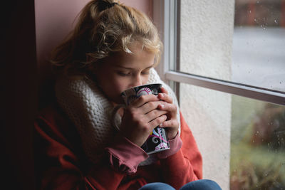 Teenage girl having coffee while sitting by window at home