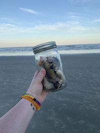 Cropped hand of woman holding glass jar on beach against sky