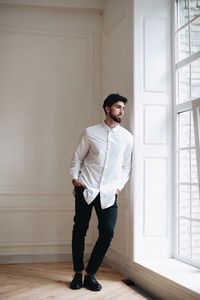 Thoughtful young man with hands in pockets looking through window at home