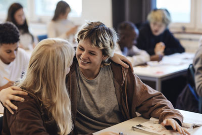 Happy female student with arm around blond girl sitting at desk in classroom