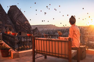 Woman sitting in balcony while looking at hot air balloons during sunset