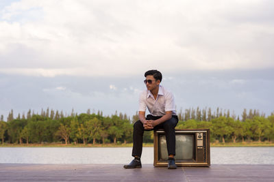 Teenage boy sitting on television set at lakeshore against sky