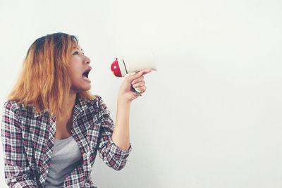 Young woman shouting on megaphone while sitting against white wall