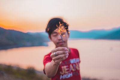 Man holding lit sparkler against lake during sunset