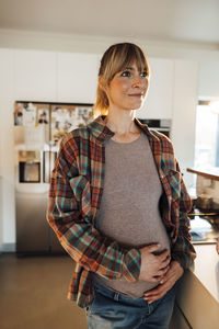 Contemplative pregnant woman with hands on stomach standing at home