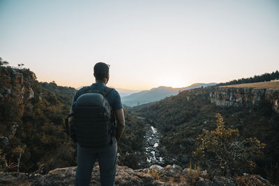 Rear view of man with backpack standing on mountain against clear sky during sunset