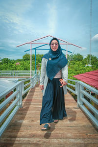 Portrait of young woman standing on footbridge against sky