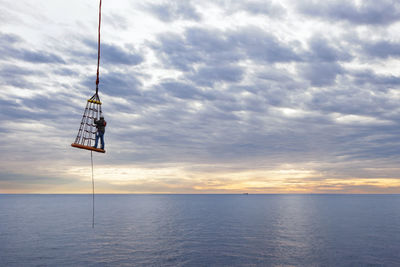 Worker lifted by offshore crane over sea against cloudy sky