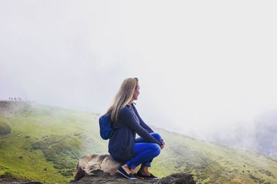 Side view of young woman sitting on rock and looking at view during foggy weather