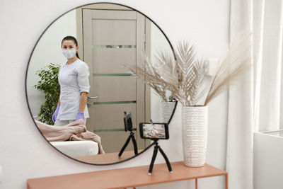 Cosmetologist in doctor's office, view though the mirror and smartphone streaming beauty procedure