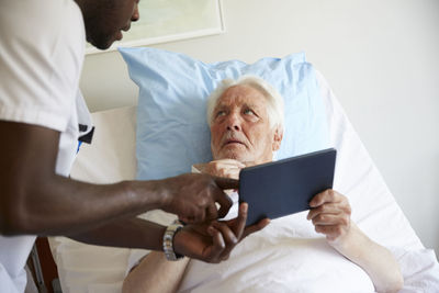Senior man looking at male nurse while using digital tablet on bed in hospital ward
