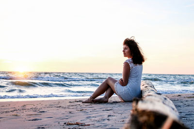 Young woman sitting on shore at beach against sky during sunset