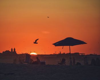 Silhouette birds flying over beach against orange sky
