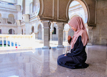 Full length of woman in hijab holding counting rosary beads while praying at mosque