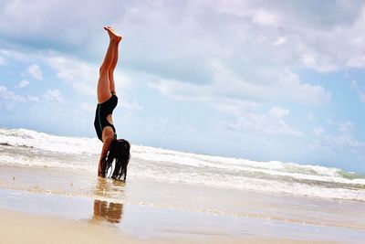 Young woman doing handstand on sea shore against cloudy sky