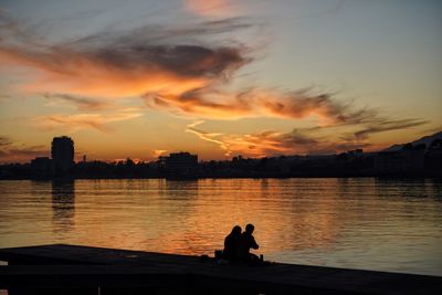 Silhouette people sitting by lake against orange sky