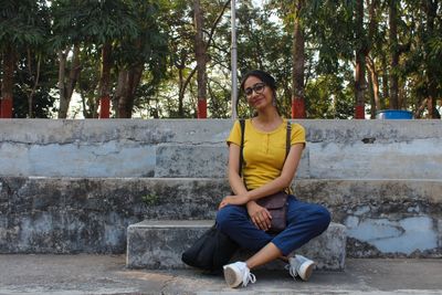 Portrait of young woman sitting on retaining wall against trees