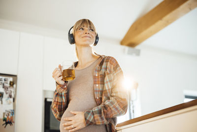 Smiling pregnant woman with headphones holding tea mug at home