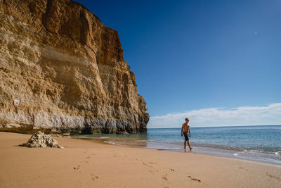 Shirtless man walking on shore at beach against blue sky