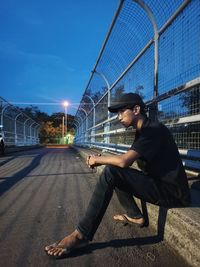 Side view of young man sitting on bridge against sky