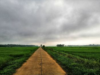 Dirt road passing through field against cloudy sky