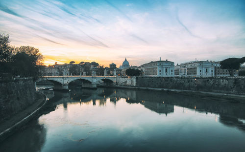 Bridge over river by buildings against sky during sunset