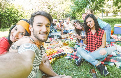 Portrait of man taking selfie with friends while siting on grass in picnic