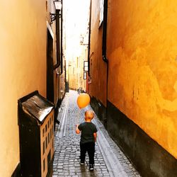 Rear view of boy with balloon walking on alley amidst buildings