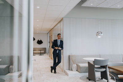 Full length of smiling young businessman with smart phone leaning on wall at corporate office