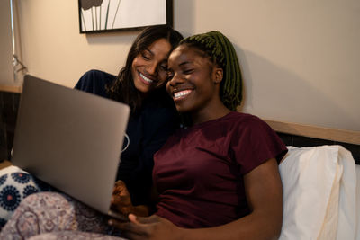 Smiling multiracial women sitting on bed and watching funny movie on netbook while relaxing together in modern bedroom at home