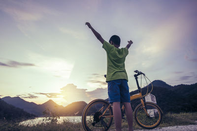 Boy standing with arms raised by bicycle against sky during sunset