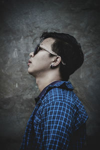 Side view of young man wearing sunglasses and blue shirt standing against wall